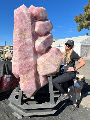 The owner, Kylee, with blonde hair and fair skin, wearing black leggings, a tee shirt, and a baseball cap. She is standing beside a huge rose quartz display piece outdoors at a gem show.