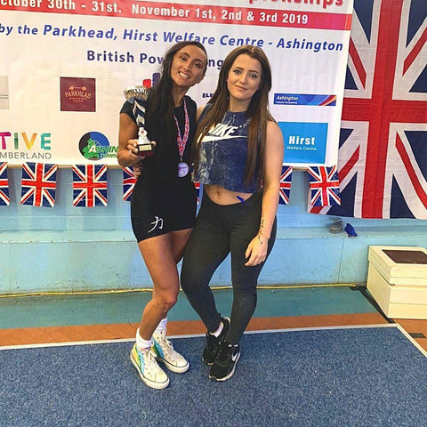 Two girls, one on the left holding a Powerlifting trophy.