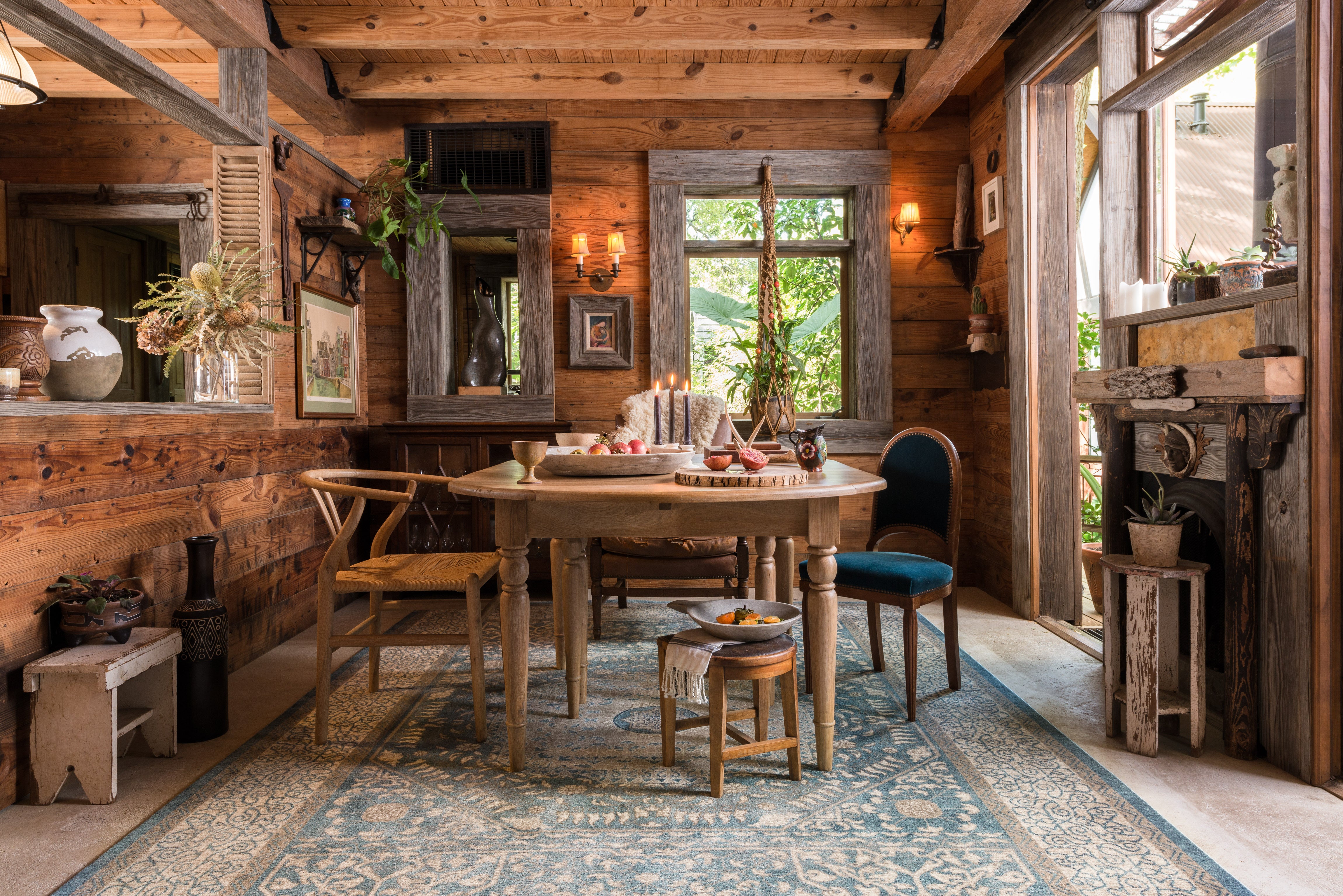 a cabin dining room with wooden walls and ceiling beams. There is a wooden table with four chairs and a blue traditional rug underneath.
