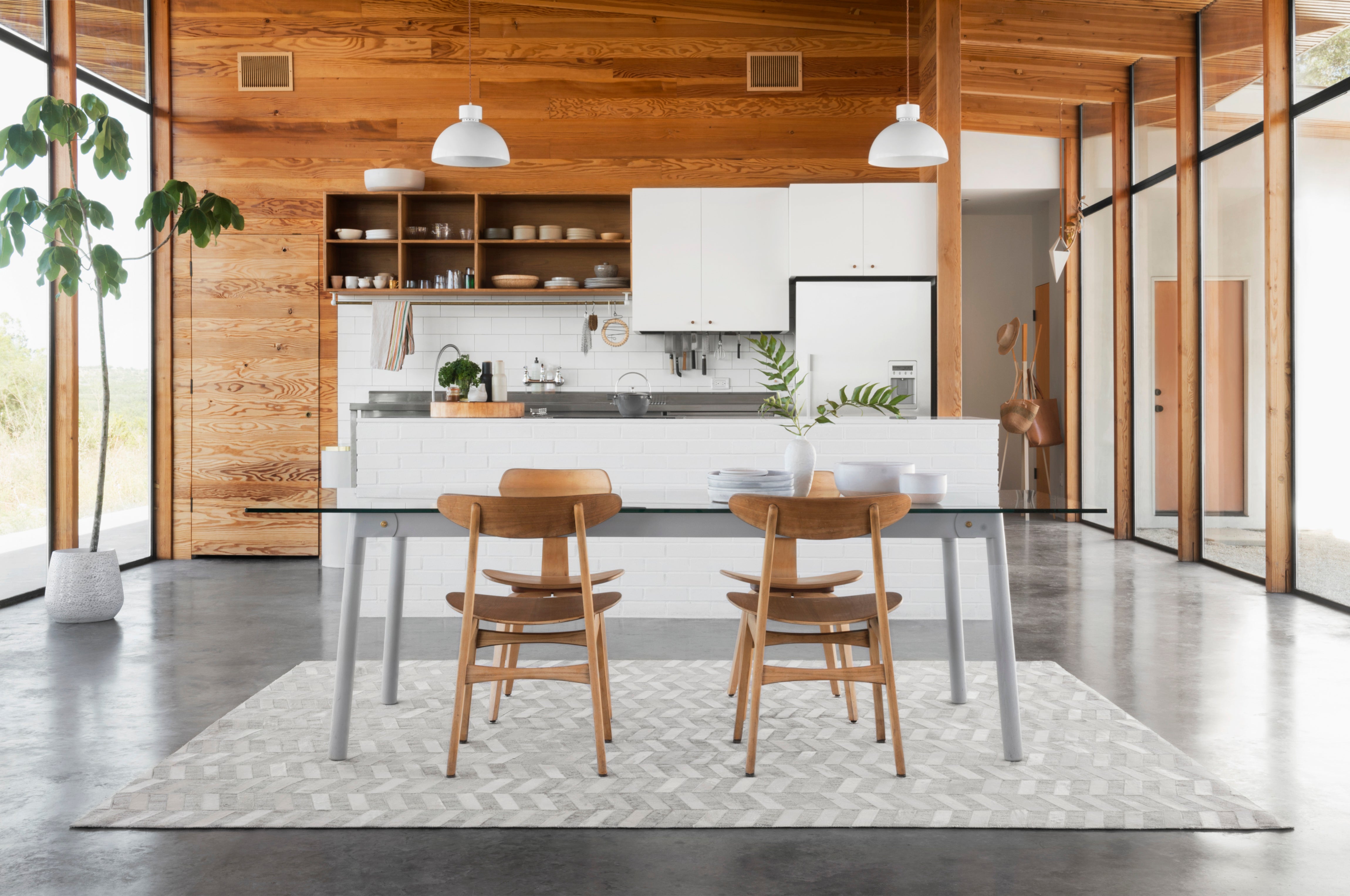 a modern table is in the foreground with four wooden chairs atop a leather, grey chevron patterned rug. In the background is a modern white kitchen with wooden lined walls and ceilings. There is a fiddle fig leaf tree in the corner and floor to ceiling windows on the right.