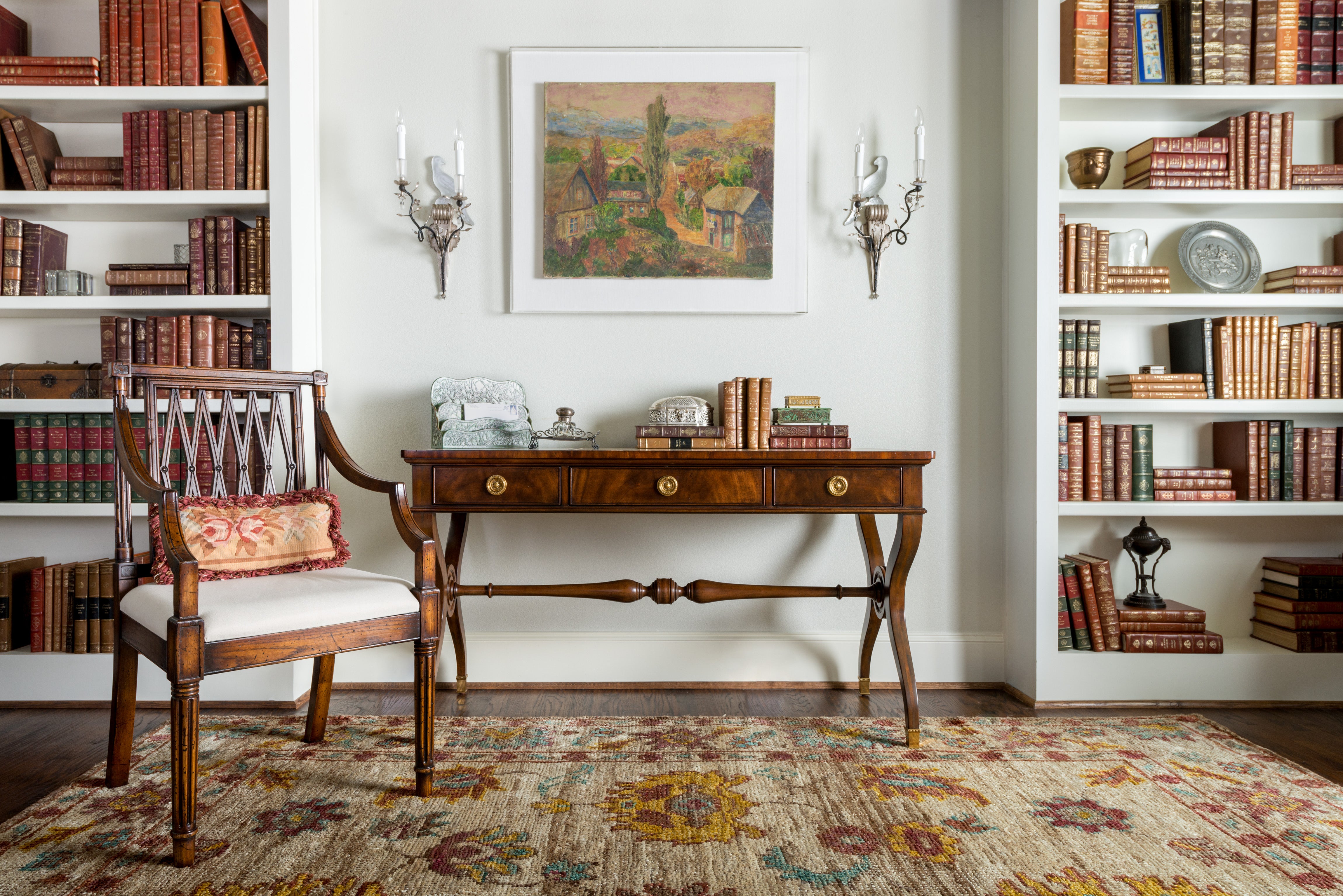 a traditional foyer table sits against a white wall with two candle sconces on either side and a traditional painting above. There are two white book shelves full of books and decorations on either side. The rug on the floor is ivory and traditional. There is one wooden chair on the left with a small pillow. 
