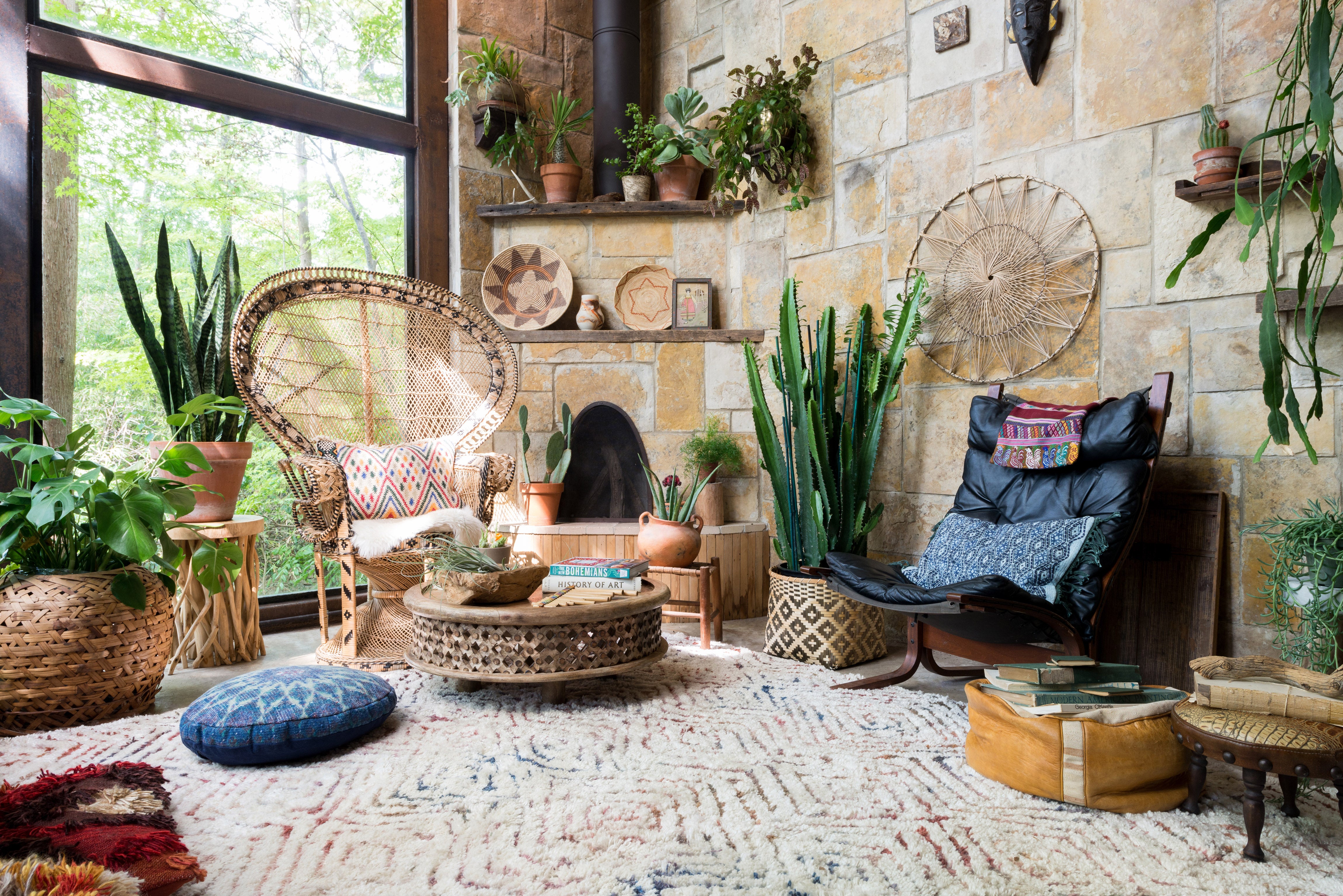 a white shag rug with multi color diamond pattern on the floor. A rattan chair and indonesian coffee table are the two main furniture pieces. The walls are stone and there is a fireplace in the corner. There are many cacti and succulent plants decorating the floor and mantle. 
