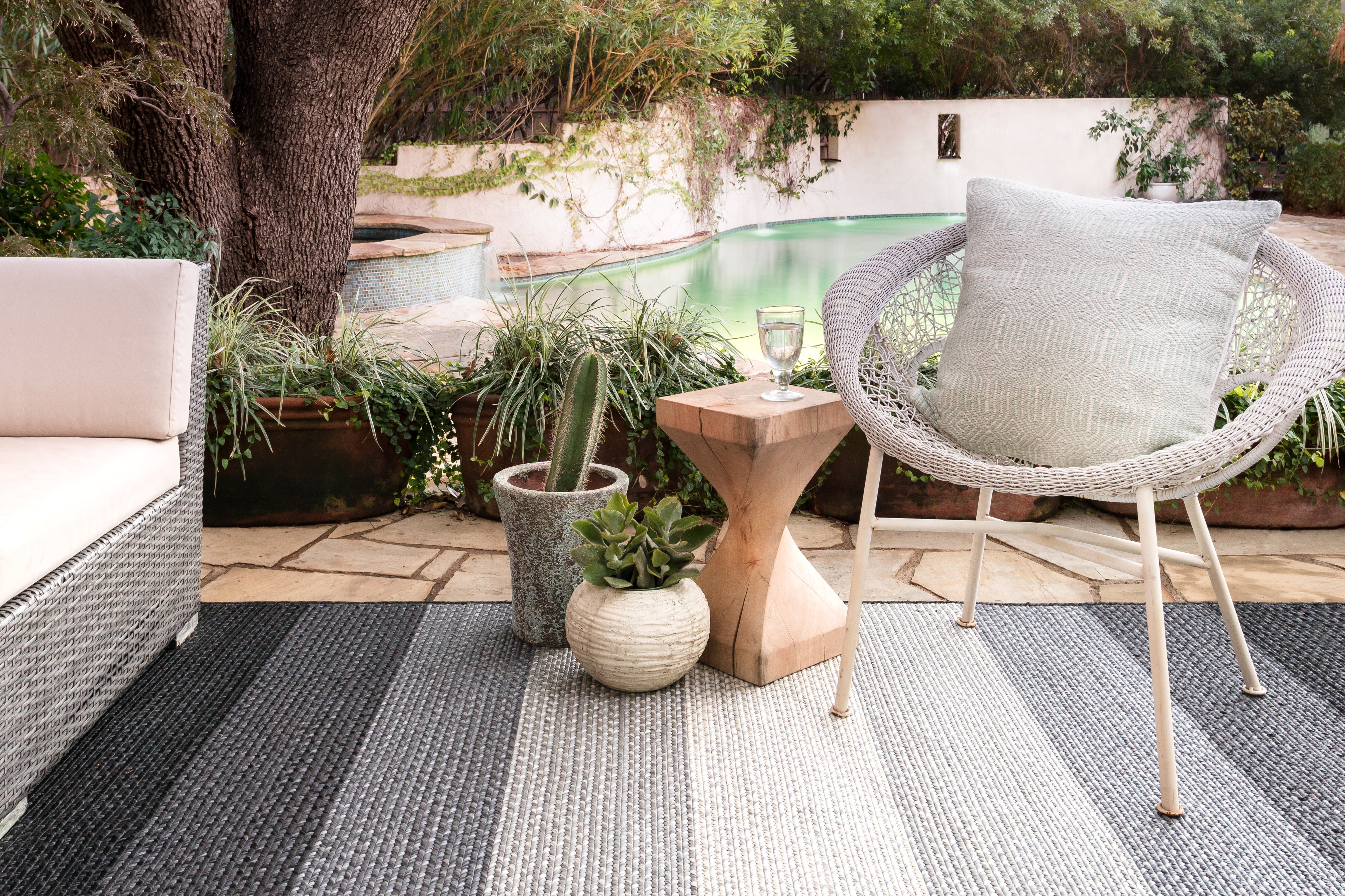 a grey and white striped outdoor area rug is opened on a patio with a pool in the background. A simple white chair and sofa sit on top with a small wooden table on the side. 