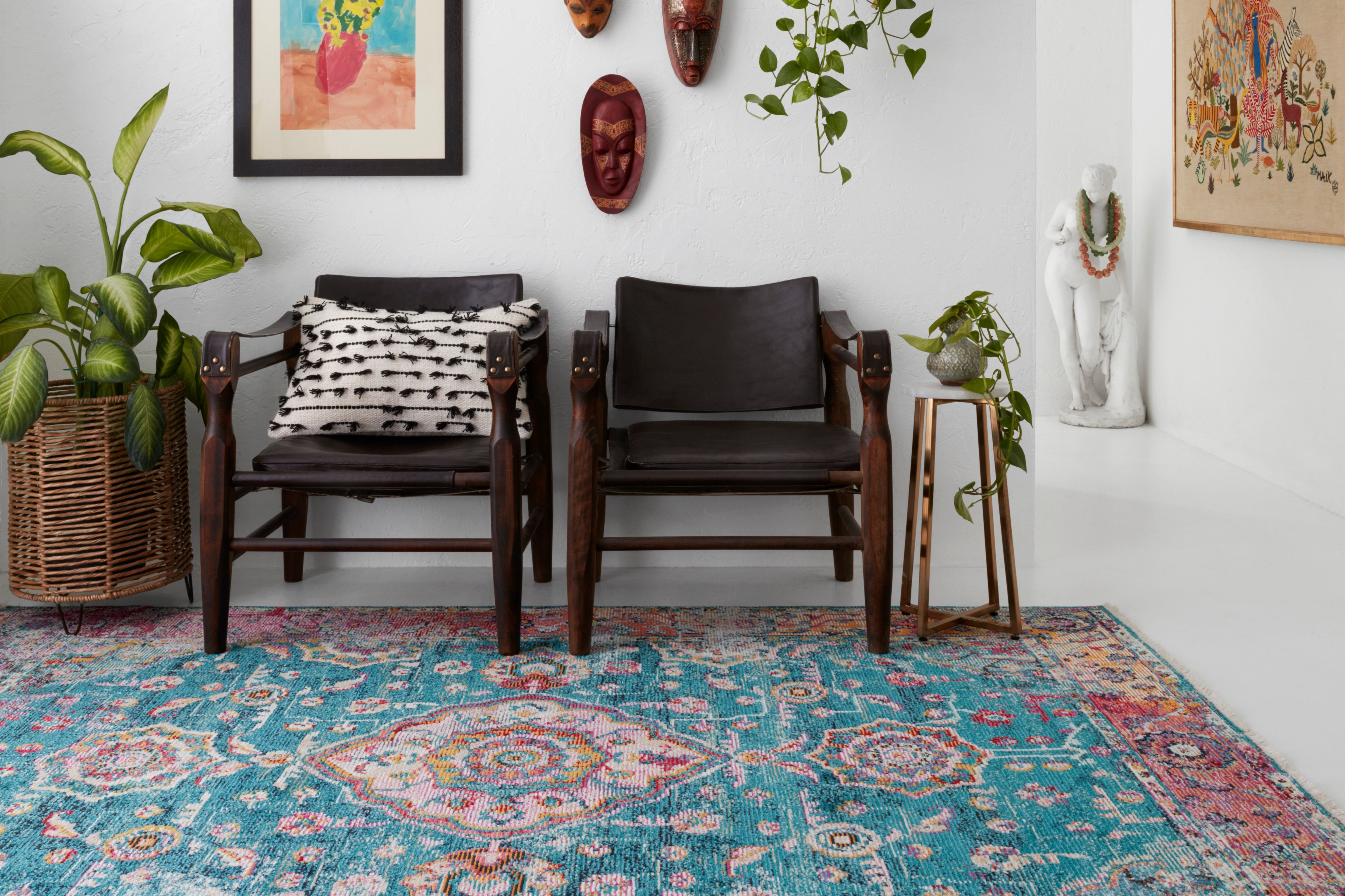 a bright blue and pink traditional rug is on the floor of an all white room. tribal wooden carved masks and paintings hang on the walls. There are two leather modern chairs against the walls. 