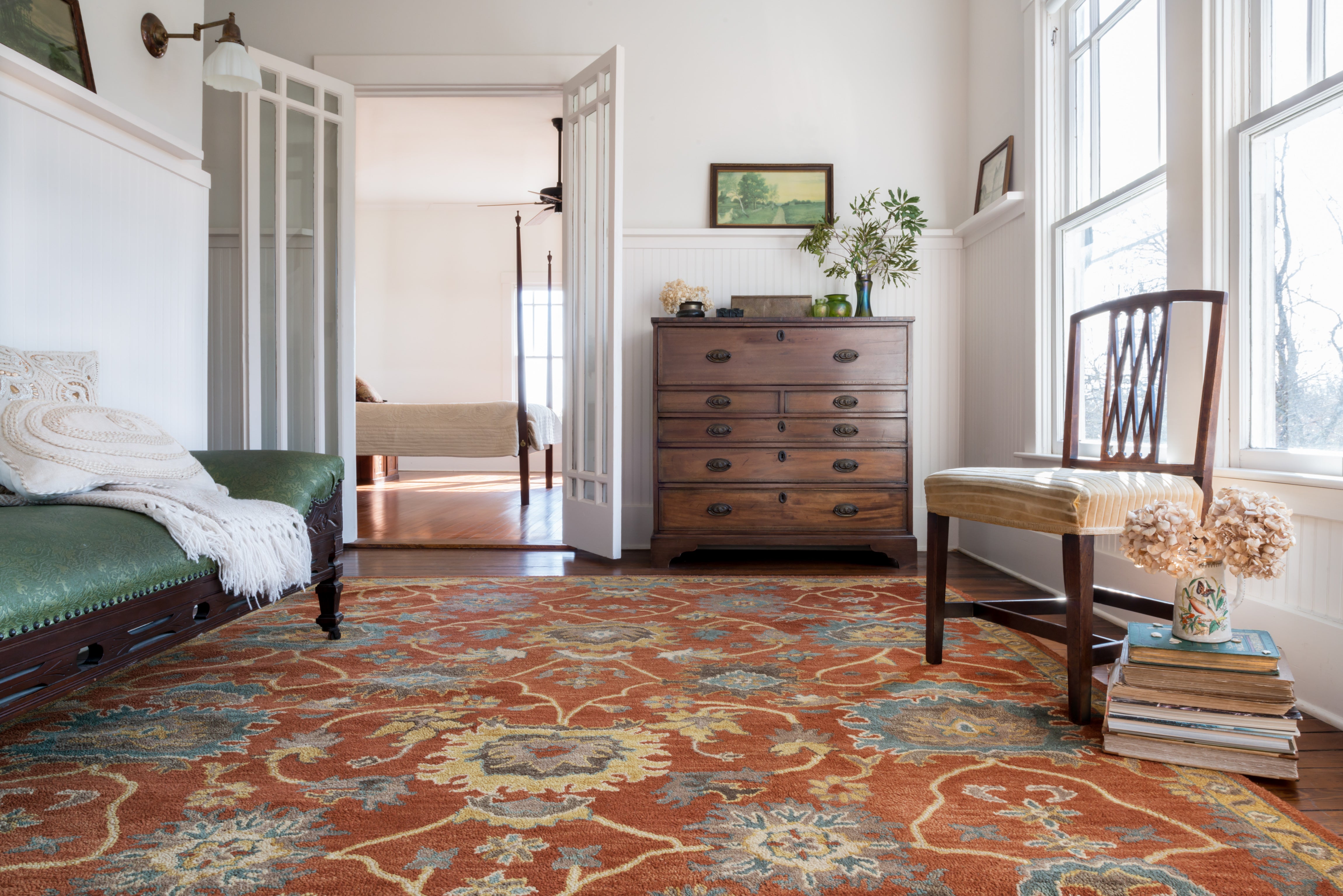 a rust colored traditional rug on the floor of a farmhouse looking bedroom. An antique dresser is against the wall and a green settee is on the left. The room is painted white. 