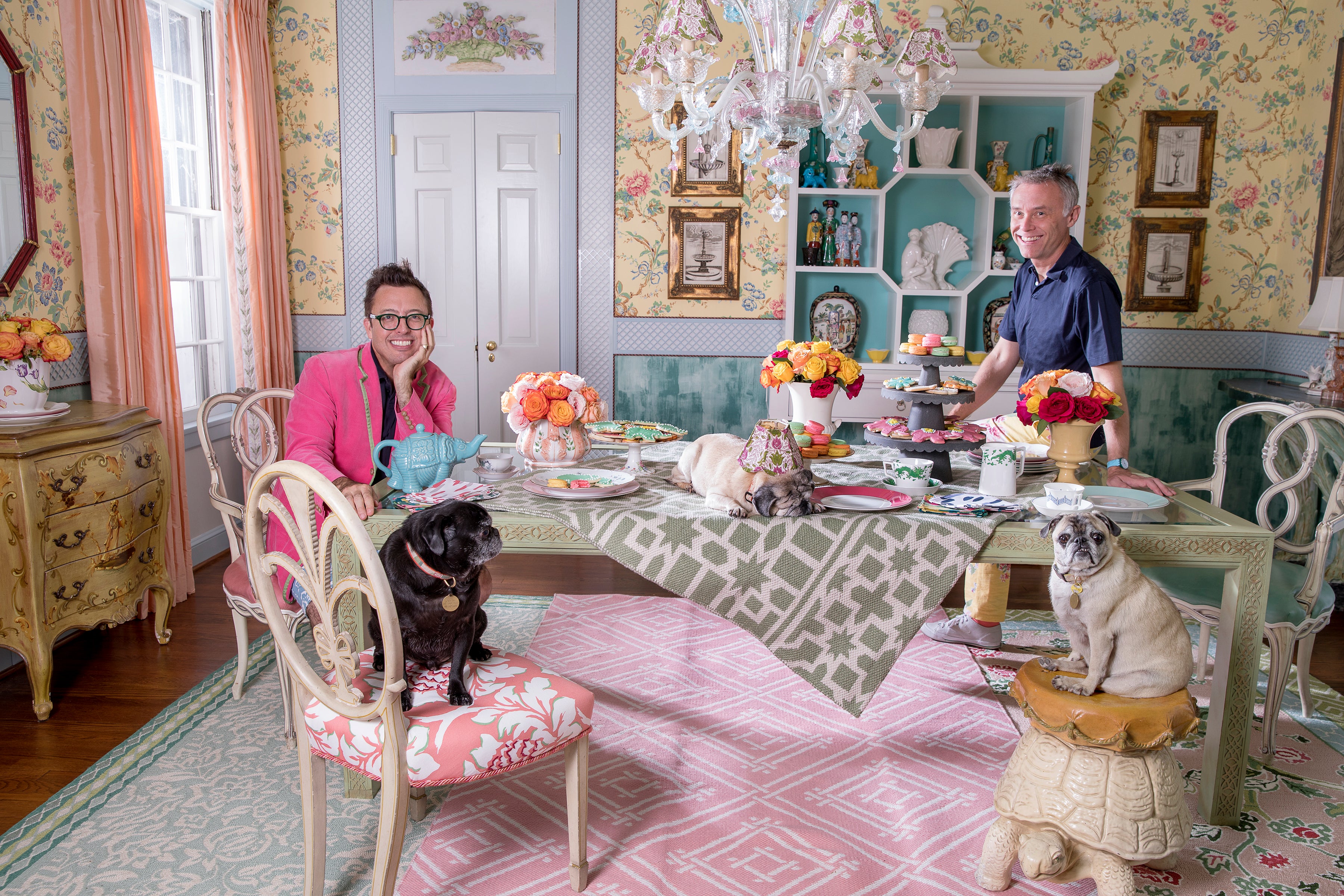 a yellow floral wallpapered dining room with a crystal chandelier hanging from the ceiling. A large table with two pug dogs sitting in the foreground and two men seated in the other two chairs. The decor is french provencal with a southern feel. There are antique furniture pieces against the walls such as china cabinets and buffet tables. The area rugs are pastel greens, pinks and yellow floral patterns.