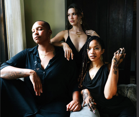 Three inclusive models lounge on a couch wearing black clothing and several Rael Cohen Jewelry pieces in gold