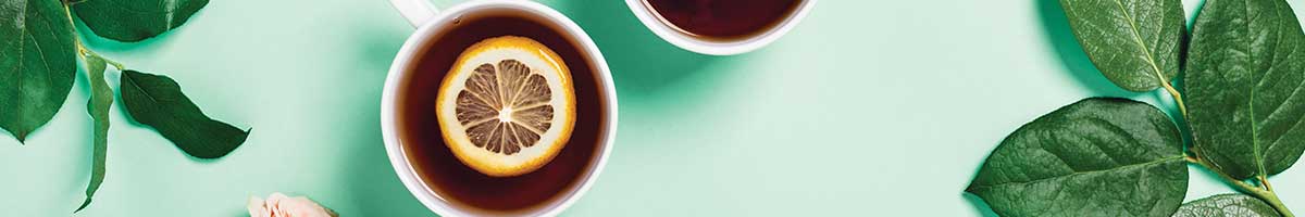 Cups of tea with lemon slices in middle of cups and green sprigs lie around the tea cups.