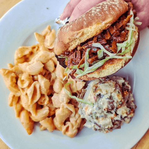 A hearty vegan BBQ sandwich with shredded lettuce, paired with creamy pasta and a side of potato salad, creating a delicious and satisfying option for vegan snacks recipes.