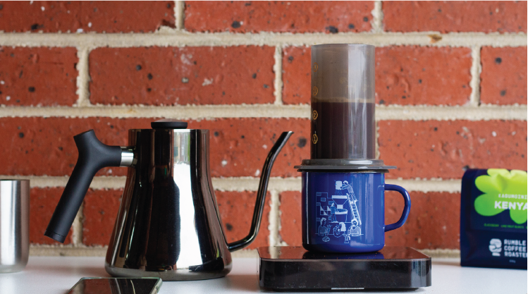 Image of Aeropress on top of a Rumble Coffee roasters enamel mug, alongside Stagg gooseneck kettle, acacia lunar scales, and a bag of Rumble Coffee Roasters single origin