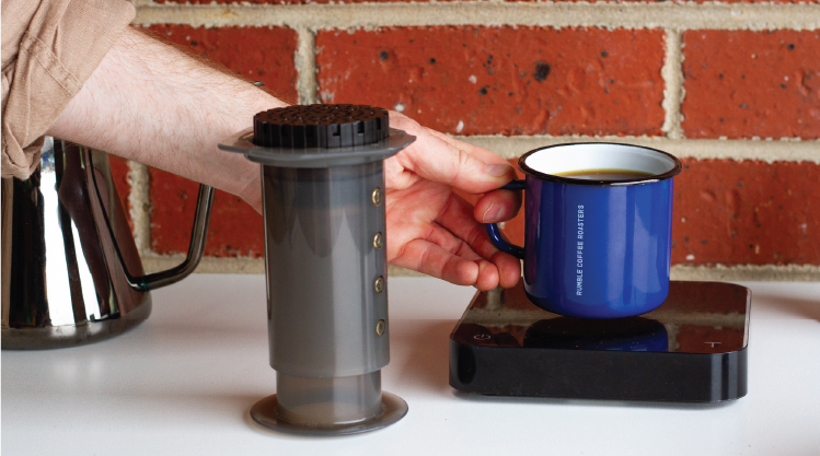 Hand reaching to grab enamel mug full of Rumble coffee, off scales. Aeropress in foreground, and filter kettle in background.