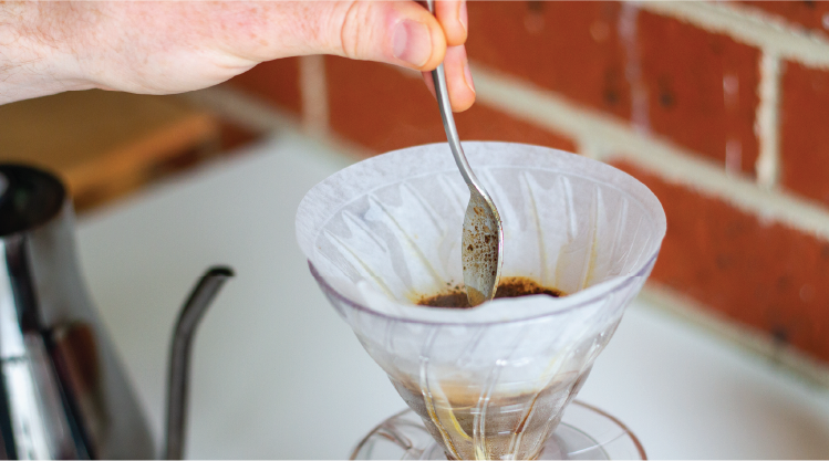 Close up of hand stirring coffee grinds in hario v60 pour over cone. Kettle visible in background