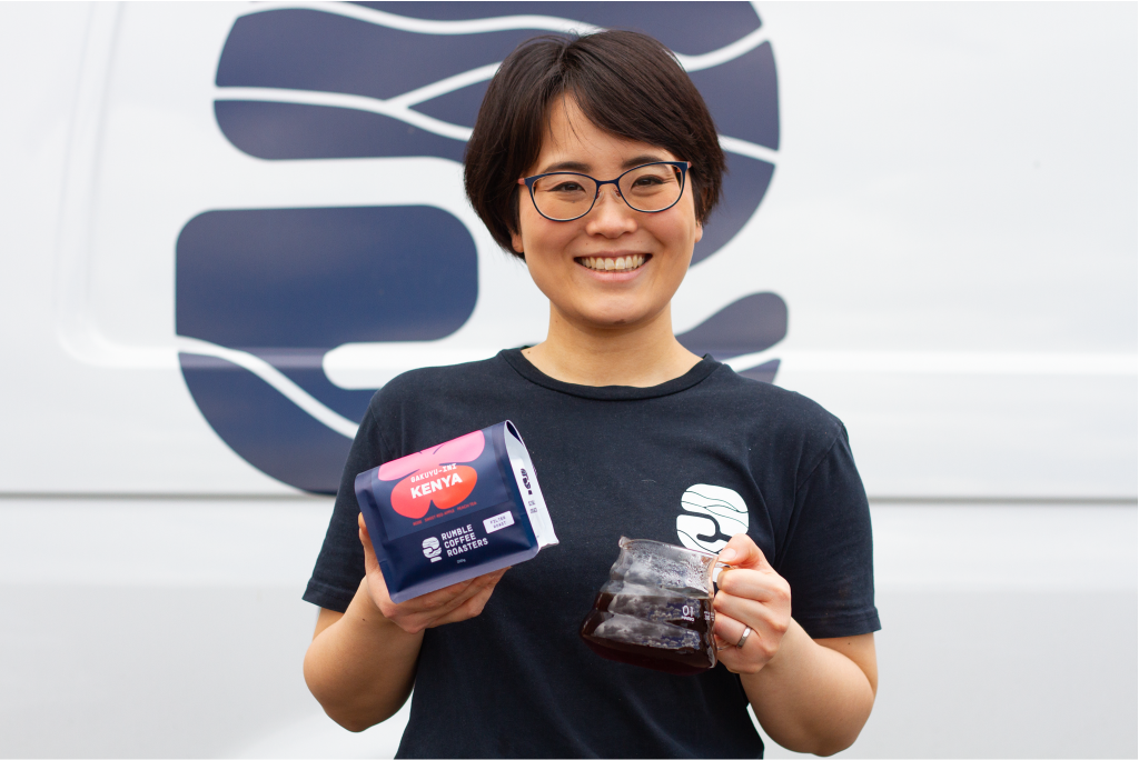 Natsumi standing in front of Rumble Coffee van, with bag of Kenya Gakuyu-ini single origin in one hand, and a carafe of filter coffee in the other.