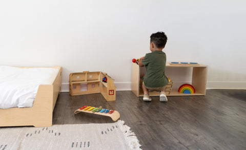 toddler boy leaning down to play at a low Montessori shelf next to a floor bed