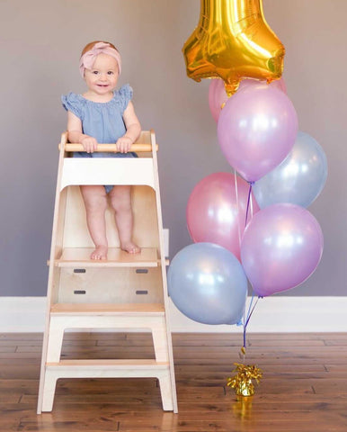 a little girl stands in a toddler learning tower with a bunch of balloons