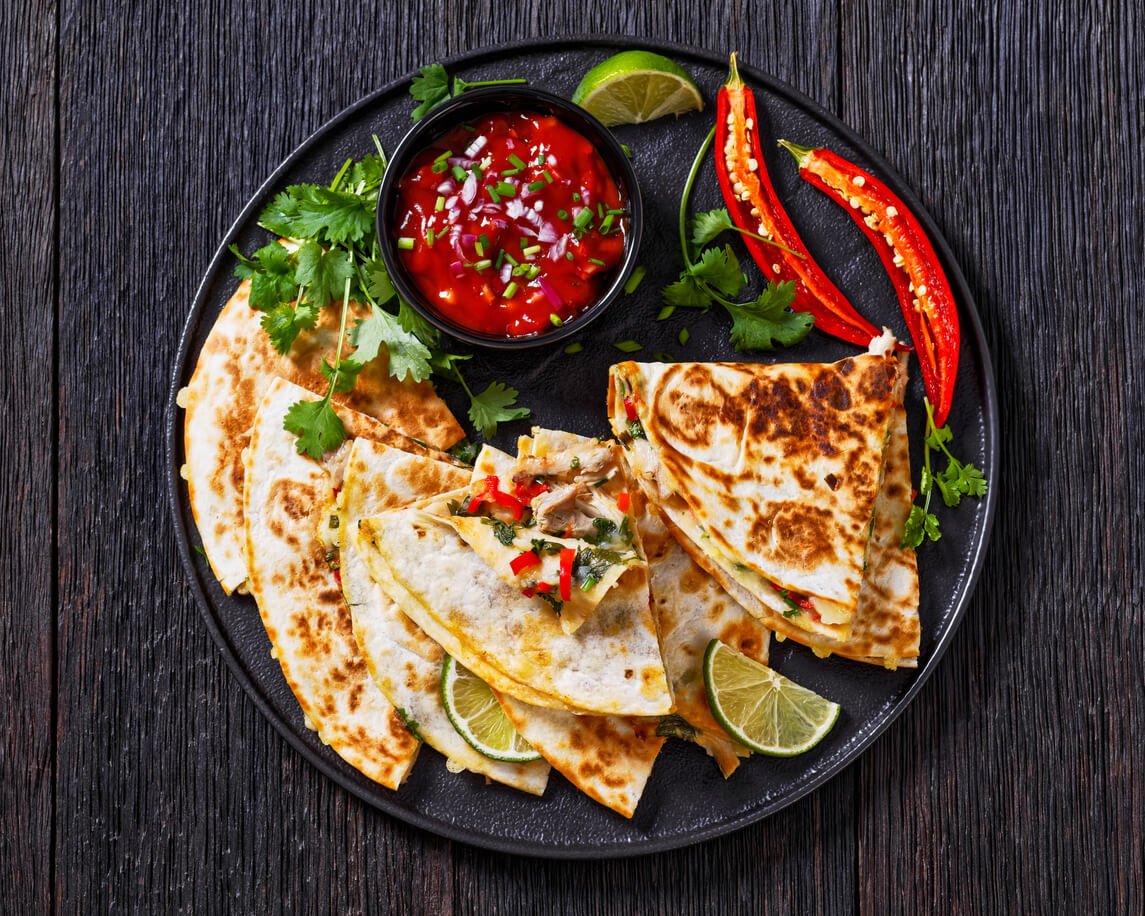 An image of quesadilla with chicken, green onion, cheese, chili pepper, coriander leaves, filling and tomato salsa served on a black plate on a dark wood table.