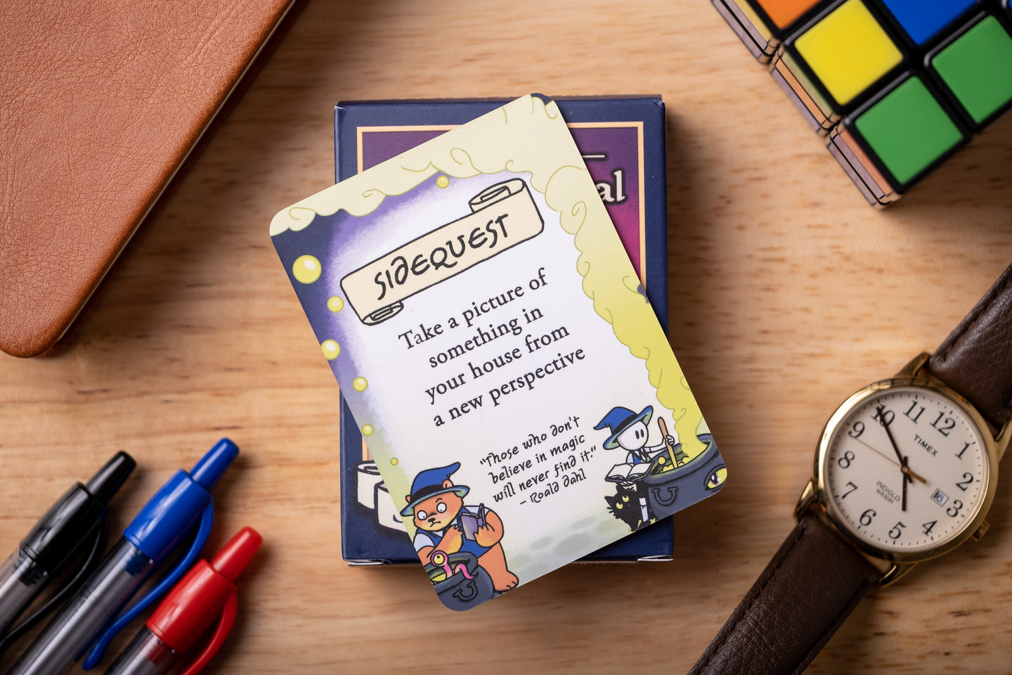 A card with a task prompt on a wooden surface surrounded by pens, a wallet, a Rubik's cube, and a watch.