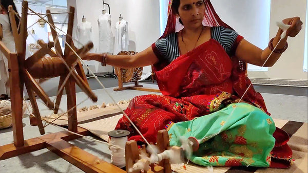 A Rajasthani women clad in bandhini saree sitting on ground and spinning cotton yarn (soot) on a Gandhi Charkha (Indian Spinning Wheel)
