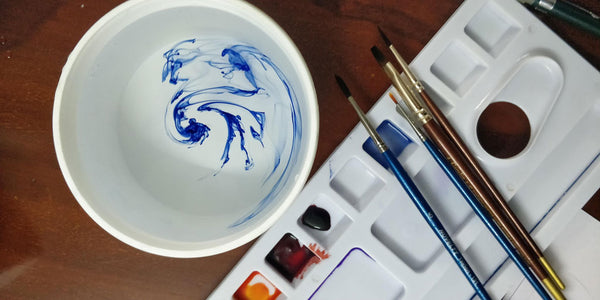a photograph of water colors in a pallete, brushes and water in a white cup from top by Rameshwari