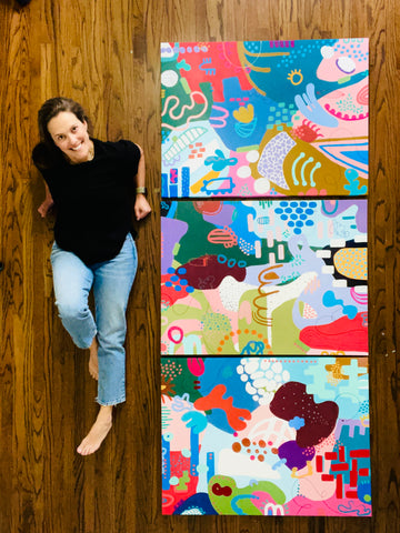 artist on the ground with three large abstract art canvases