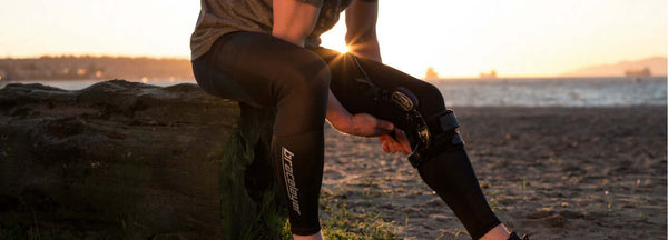 A man on a beach putting on a knee brace over his Bracelayer pants, which provide great meniscus tear knee support!