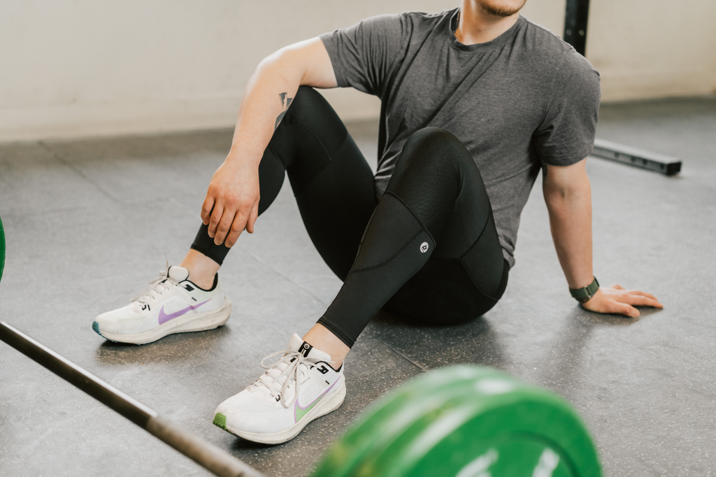 A man resting between sets with a large barbell in the foreground. He is sitting with his knees up, resting one arm on his left knee and leaning back on his right arm. He is wearing Bracelayer's KS1 knee brace compression pants for added knee support and stability during his workout.