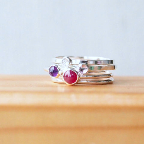 Five stacking rings with a mix of gems on a wooden background, the gems are ruby, Amethyst, and white topaz in a mix of sizes and are sterling silver. Handmade by maram jewellery in Scotland