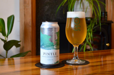 A can of Burnt Mill Pintle pale ale next to a Caps and Taps tulip glass that it has been poured into on a coffee table. The beer is a clear amber colour.