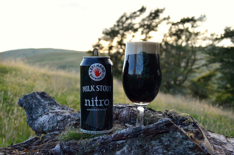 A can of Left Hand Brewing Co Milk Stout Nitro next to the Caps and Taps tulip glass it has been poured into, both on a log. The beer is a very dark brown with a thick creamy head