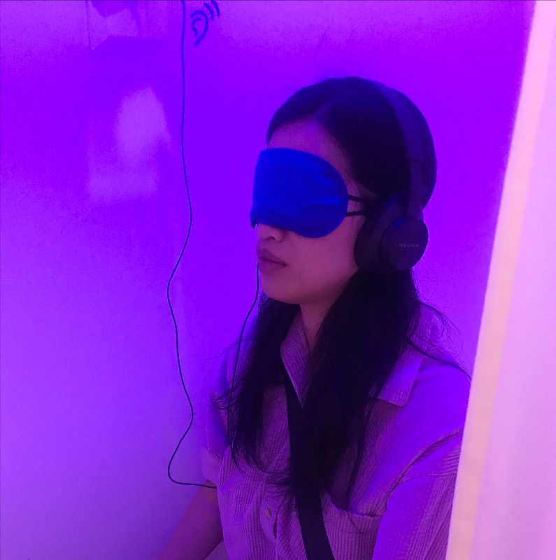 A viewer sits in one of the makeshift sound booths, surrounded by soothing blue and pink lighting. They have on a blindfold and headphones to listen to the CHKRA album.
