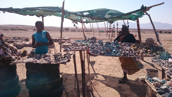 In the Namibian desert under a shade cloth, behind tables full of rocks, two traders await customers