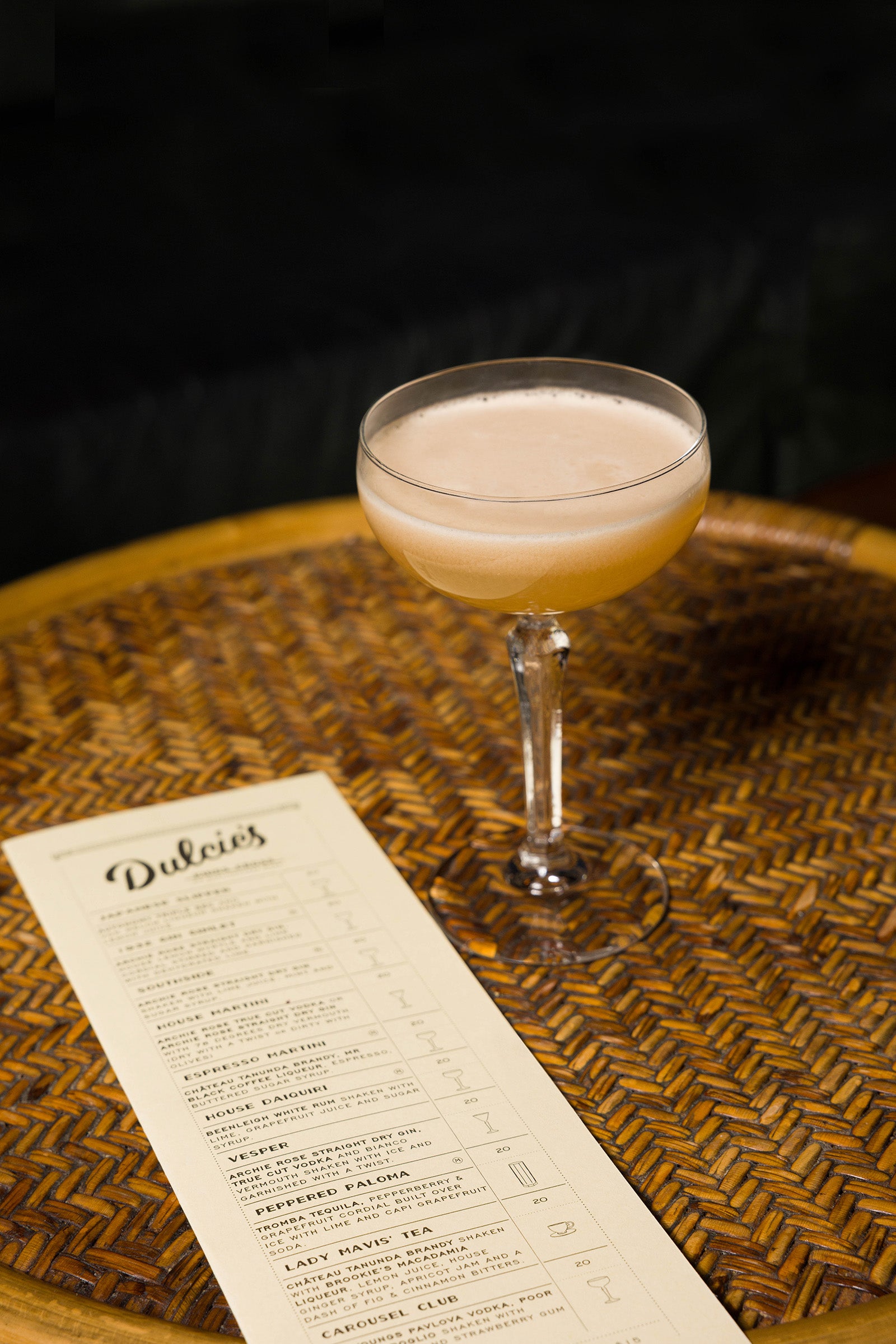 A whisky sour cocktail with white foam top in glassware next to the Dulcies bar menu