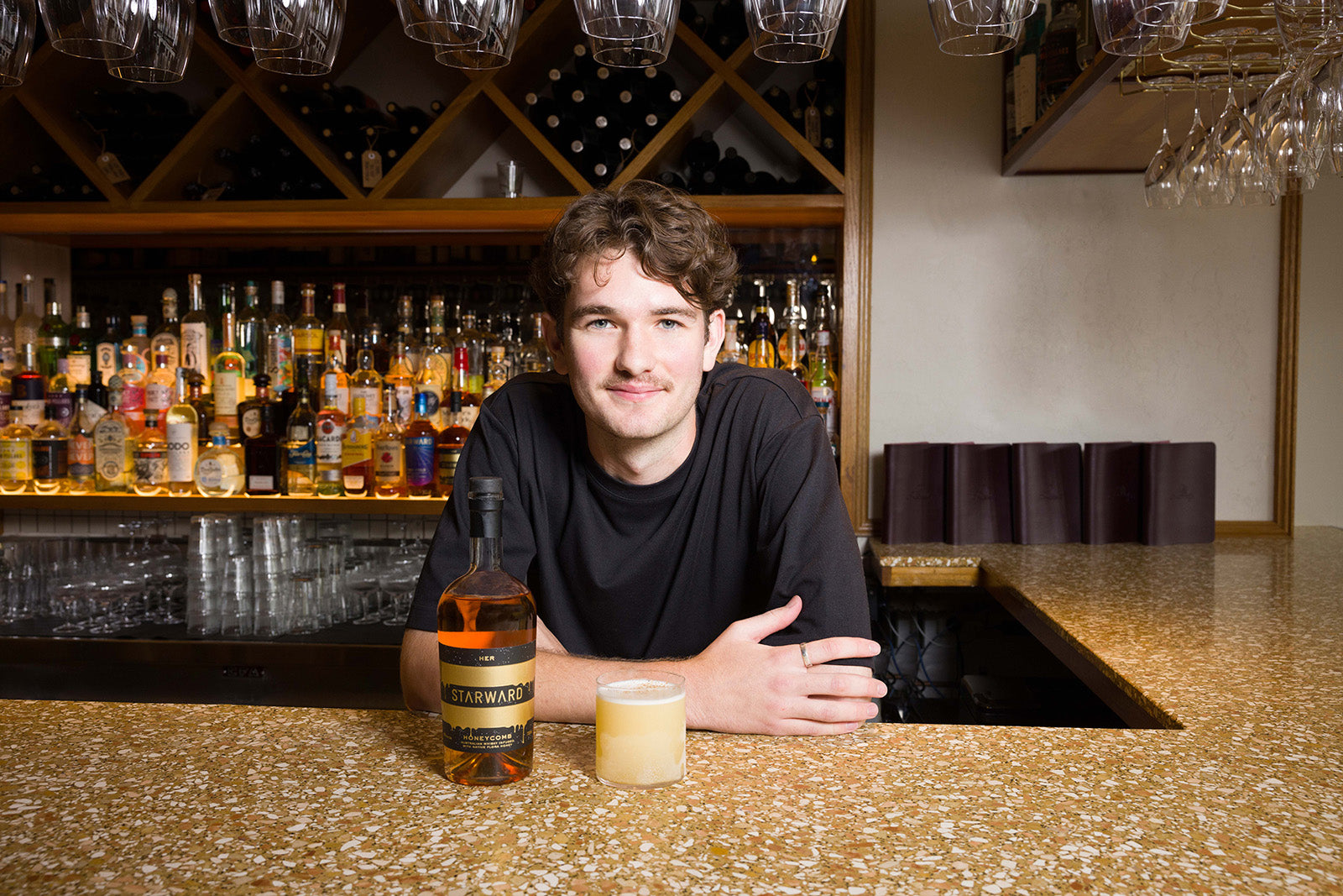 A man leaning across a bar with a cocktail he has just made from HER Honeycomb Starward Whisky