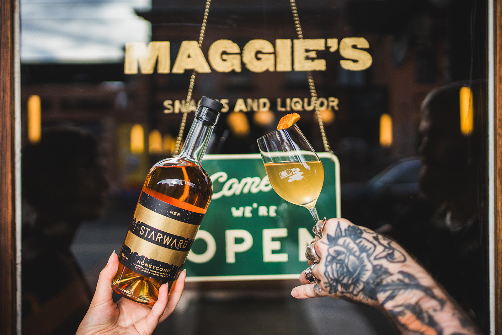 A bottle of whisky and class of whisky coloured liquid being cheers in front of a bar