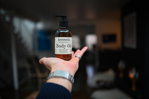 Aeemelia Body Oil, a brown amber pump bottle with an off white label being held by an out stretched hand. 