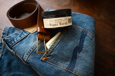 Aeemelia Sugar Scrub brown amber container with off white label on a pair of jeans next to a worn leather belt