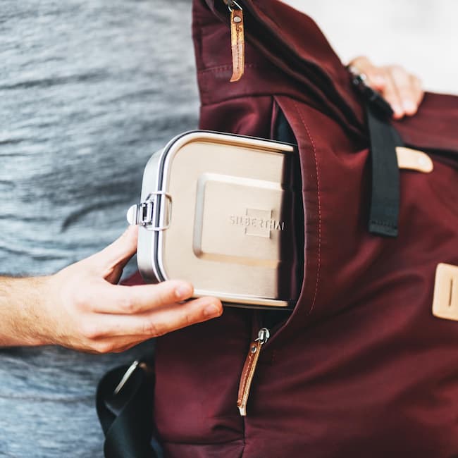 Die Edelstahl Brotdose wird in einem Rucksack gesteckt. Text Ideal für Picknick, Schule oder Büro