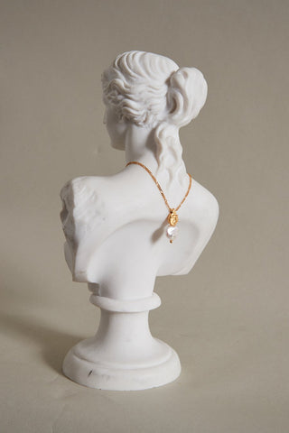 A ceramic bust faces away, with a decadent twisted gold and pearl necklace dangling down the back of its neck