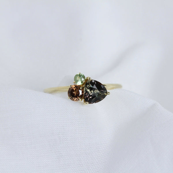 A gold cluster ring featuring a multi-coloured sapphire alongside brown and green gems, on white fabric