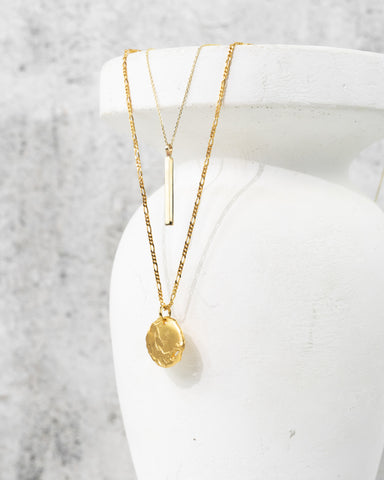 Two gold necklaces hang from a white vase. One necklace is a gold vertical bar, the second necklace features a rough gold coin. 