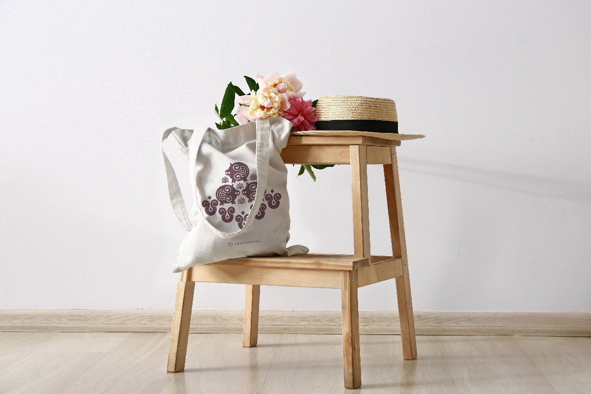 Circe tote bag on a chair with summer flowers and a straw hat.