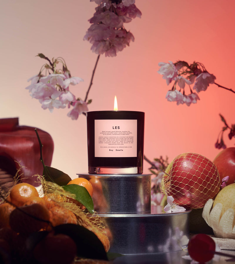 Editorial of Les with a lit wick on top of a silver metal base amongst fruit and a floral arrangement against a pink orange background. 