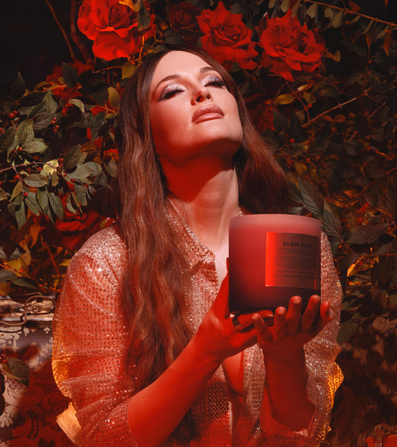 Editorial shot of a women holding a large slow burn candle with a look of bliss on her face, she is wearing a metallic see through flowy shirt, has metallic silver eye shadow and long wavy brown hair. Her head is faced up towards the sky and behind her is a large rose bush. She is bottom lit with a red glow.