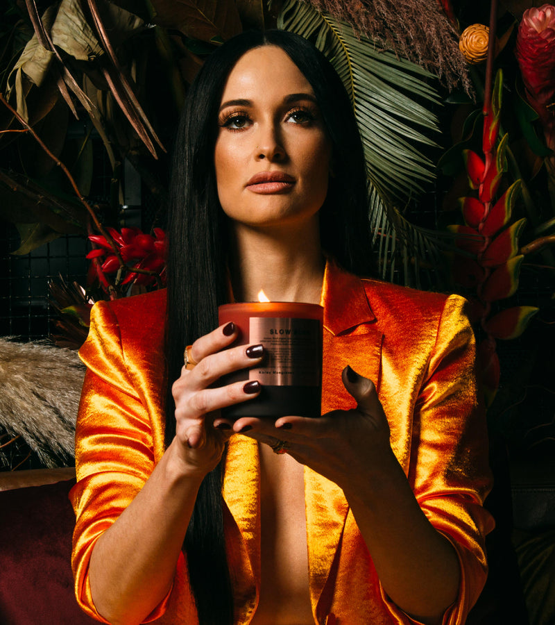 Editorial shot of slow burn held by a powerful looking women in a 3/4th shot with long black hair, a orange velvet blazer and deep red nails holding the candle in front of her while amongst a forest background.
