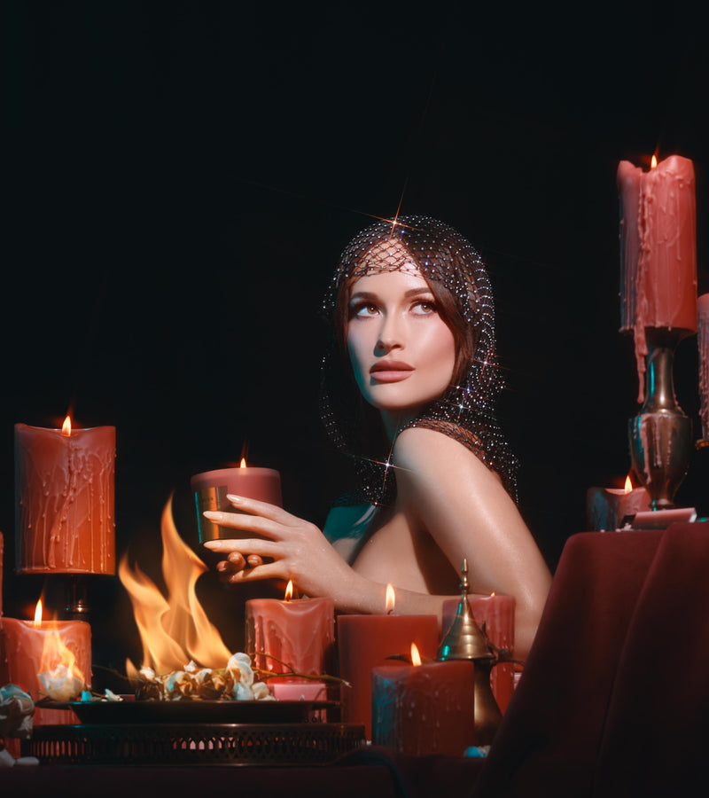 Editorial shot of a women with a light chain mail vail holding slow burn candle while surrounded by other dripping red candles and a plate of flowers on fire against a black background.