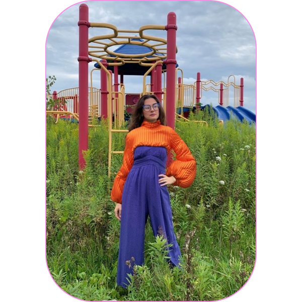 woman standing in front of an overgrown abandoned playground wearing a blue jumpsuit and a cropped orange digital puffer jacket