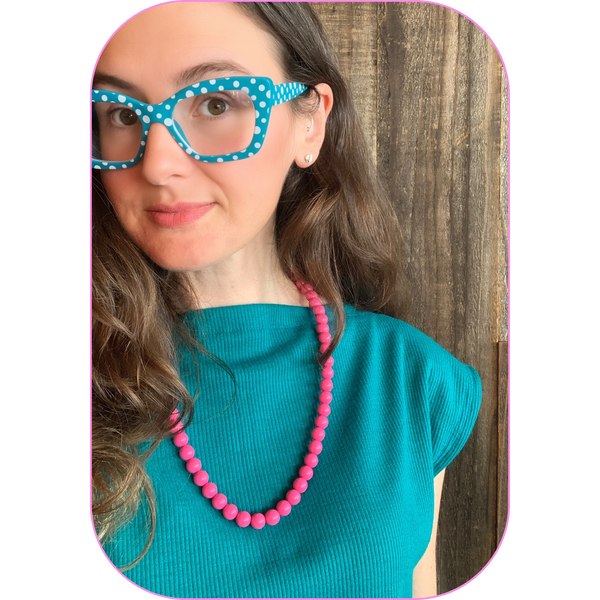 woman wearing a teal boat neck top and vintage pink beaded necklace standing in front of a wood paneled wall