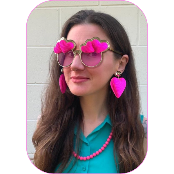 close-up shot of a woman wearing a turquoise sleeveless button-down shirt, a pink beaded necklace, and digital sunglasses adorned with pink rhinestone hearts with matching earrings