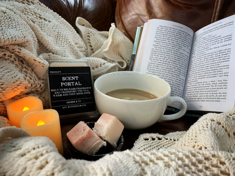Image shows the Cosy Book Nook Scent Portal wax melt displayed with a cup of coffee, candles and an open book to create a cosy place to read.