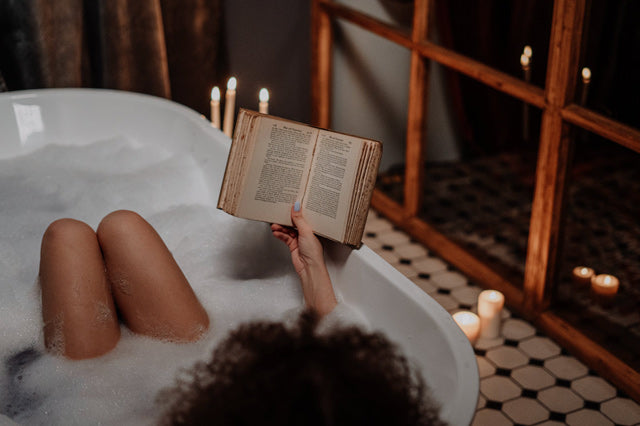 woman relaxing in bubble bath, reading a book surrounded by candles